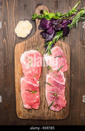 Zwei appetitliche rohes Schweinefleisch Steaks, auf einer hölzernen Schneidebrett, mit Knoblauch und verschiedene frische Kräuter und Gewürze, Ansicht von oben Stockfoto