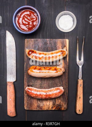 Gebratene Würstchen auf einem vintage Schneidebrett mit Messer und Gabel für Fleisch, Ketchup und Salz, Ansicht von oben Stockfoto