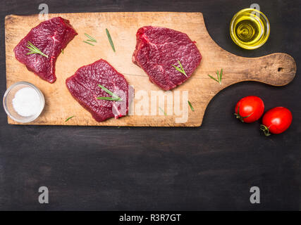 Appetitlich rohes Rindfleisch, auf einem vintage Schneidbrett, mit Rosmarin, Tomaten, Butter und Gewürze, Grenze, Platz für Text auf rustikalen Holzmöbeln Hintergrund Stockfoto