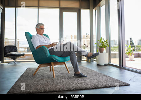 Reifer Mann im Sessel zu Hause sitzen mit Tablet Stockfoto