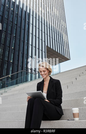 Blond Geschäftsfrau mit Kaffee zu sitzen auf der Treppe vor der modernen mit Tablet Bürogebäude gehen Stockfoto