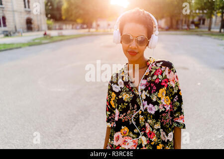 Modische junge Frau mit Sonnenbrille und Kopfhörer im Freien bei Sonnenuntergang Stockfoto