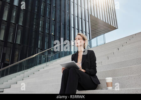 Blond Geschäftsfrau mit Tablet und Kaffee zu sitzen auf der Treppe vor der modernen Bürogebäude gehen Stockfoto