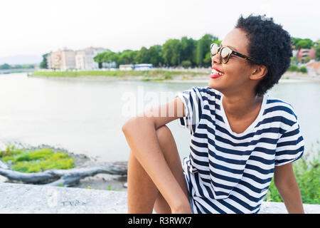 Lächelnde junge Frau mit Sonnenbrille am Fluss sitzen Stockfoto