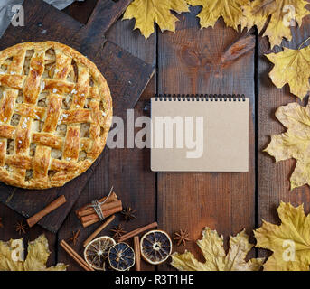 Gebackene ganze Runde Apfelkuchen auf braunem Holz- Board, Blätterteig, und eine leere Notepad öffnen mit braunen Seiten für die Rezeptur Stockfoto