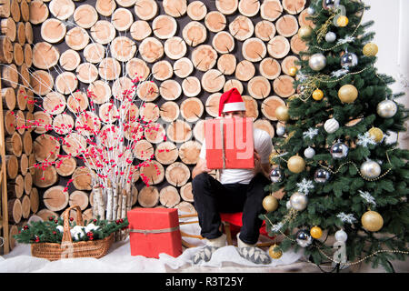 Mann in santa hut Peep über den roten Kasten auf Schlitten am Weihnachtsbaum, Korb, Red berry Bush auf Schnee Dekorationen. Boxing Day Konzept. Stockfoto