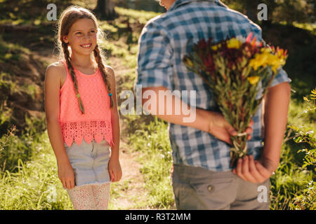 Junge stand vor seiner Freundin in einem Park mit Blumen hinter ihm. Junge Ausdruck seiner Liebe zu einem Mädchen versteckt sich ein Haufen Blumen b Stockfoto