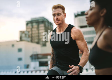 Fitness-Mann und -Frau, die auf einem Dach trainieren, mit Hochhäusern im Hintergrund. Athleten, die früh auf der Terrasse trainieren Stockfoto