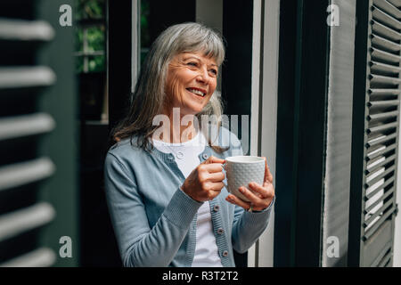 Ältere Frau, die in der Nähe der Tür, Kaffee zu trinken. Lächelnd alten Frau trinkt Kaffee mit Blick auf die Tür. Stockfoto