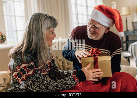 Älterer Mann, Santa cap geben ein Weihnachtsgeschenk für seine Frau. Alte Mann sitzt zu Hause mit seiner Frau, die ihr ein Geschenk. Stockfoto