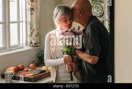 Senior Paar stehen in der Küche mit einem Blumenstrauß. Älterer Mann küsste seine Frau hielt ihre Hand zu Hause. Stockfoto