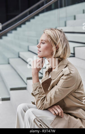 Blonde junge geschäftsfrau tragen beige Trenchcoat sitzt auf der Treppe Stockfoto