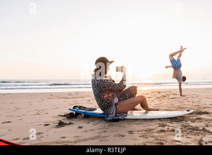 Junge Frau sitzt auf Surfboard, die Bilder des jungen Mannes, üben Handstand am Strand Stockfoto