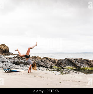 Junge Frau mit Surfbrett macht handstand am Strand Stockfoto