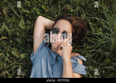 Porträt der lächelnde Frau mit Sonnenbrille im Gras liegend mit Blume im Mund Stockfoto