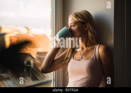 Blonde junge Frau trinkt Kaffee aus der Tasse Blick aus Fenster Stockfoto