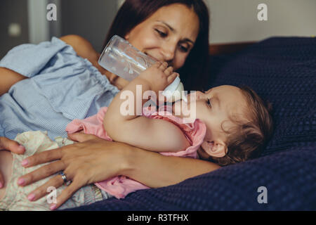 Die Mutter, die ihr Baby Mädchen trinken Milch aus der Flasche auf dem Bett Stockfoto