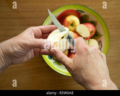 Hände einer Frau coring Eine organische honeycrisp Apple mit dem Zinn Schale mit Äpfeln auf einem hölzernen Tisch im Hintergrund. Stockfoto