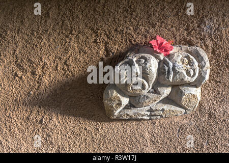 Zwei geschnitzte Gesichter auf eine sehr rauhe Wand in Bali mit einer roten Blume auf der Oberseite und dramatische Beleuchtung. Stockfoto