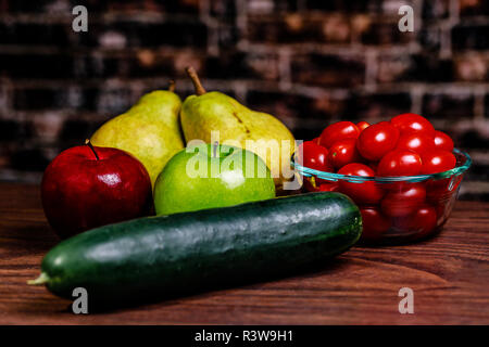 Gurken, Birnen, Äpfel und Tomaten auf einer hölzernen Oberfläche zusammengefasst und zum Schneiden bereit und gegessen oder in einer Schale verwendet werden. Der Hintergrund ist ein Vintage brick Stockfoto