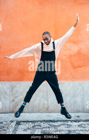 Junge schwarze Mann legere Kleidung im städtischen Hintergrund springen. Lifestyle Konzept. Tausendjährigen afrikanischen Kerl mit Latzhose im Freien