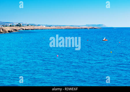 Bild der ruhigen Meer Oberfläche in der Nähe von Agia Napa, Zypern. Eine orange Tretboot im tiefen blauen Wasser, das gegen die felsige Küste und Cape Greco auf dem Hintergrund. Warm Stockfoto