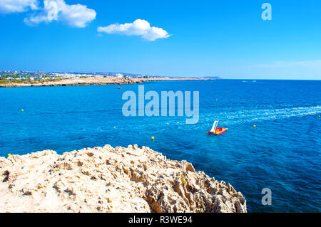 Bild der ruhigen Meer Oberfläche in der Nähe von Agia Napa, Zypern. Eine orange Tretboot im tiefen blauen Wasser, das gegen die felsige Küste und Cape Greco auf dem Hintergrund. Warm Stockfoto