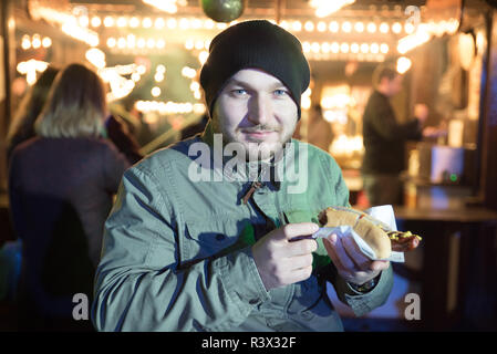 LONDON, Großbritannien - 23 November, Hyde Park 2018 Winter Wonderland traditionelle Kirmes. Mann essen Wurst vor der Garküche der Weihnachtsmarkt Winter Stockfoto