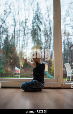 Kleine blonde Junge kniend auf einem Holzboden zu Hause vor der Tür ein Glas Terrasse mit Blick auf den Garten mit seiner Hand auf das Glas im Abendlicht. Stockfoto