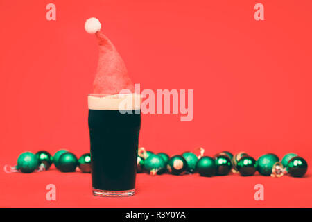 Nonik pint Glas dunkles Bier oder stout Ale mit rote Weihnachtsmütze und verschwommenes grün Christbaumkugeln im Hintergrund auf Rot Stockfoto