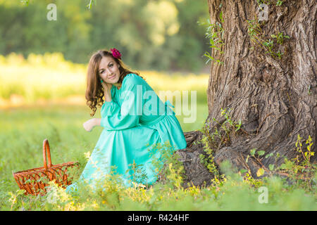 Eine Mädchen in einem langen Kleid in einer guten Stimmung kauerte in der Nähe von einem Baum im Wald Stockfoto