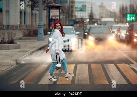 Rothaarige junge Frau in einem weißen Mantel und Blue Jeans überquert die Straße an einem fußgängerüberweg. Autos für den Hintergrund. Stockfoto