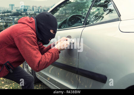 Der Mann im schwarzen Räuber Maske brechen das Auto Lock auf einer Stadt Hintergrund. Krank - beabsichtigte Dieb Kräfte, um das Fahrzeug zu verriegeln mit einem picklock. Übeltäter Stockfoto