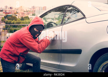 Maskierte Mann hockt und bricht ein Auto auf den Betrachter. Dieb in Hoodie und verborgene Gesicht macht versuchen, picklock das geparkte Fahrzeug zur Festsetzung Stockfoto