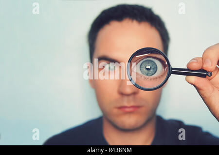 Portrait des jungen Mannes durch die Lupe auf weißem Suchen verschwommenen Hintergrund. Blick auf männliche blauen Auge durch das Objektiv. Mit Makro Auge in der Hand, die Person im Vordergrund Lupe. Informationen suchen. Stockfoto