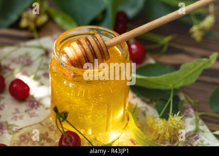Öffnen Glas Glas von flüssigen Honig und Honig Pendelarm, Bündel von Linden Blumen und rote Kirsche auf Holz- Oberfläche. Sonnenstrahl. Dunklen rustikalen Stil. Stockfoto