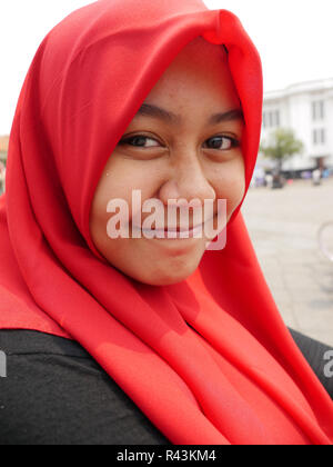 Vertikale Porträt der jungen indonesischen Mädchen mit grossen Grinsen und hellen Augen, trägt einen roten Schal und Schwarz bei Camera suchen Kleid. Stockfoto