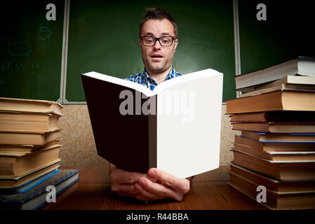 Überrascht zerzaust müde Unrasierten jungen Mann in Gläsern, ein Buch zu lesen, die mit zu Tisch Stapel der Bücher vor dem Hintergrund der Tafel. Stockfoto