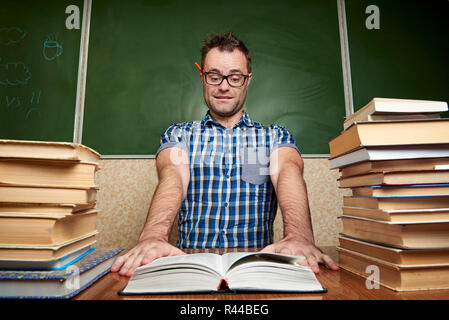 Mad zerzaust müde Unrasierten jungen Mann in Gläsern, ein Buch zu lesen, die mit zu Tisch Stapel der Bücher vor dem Hintergrund der Tafel. Stockfoto