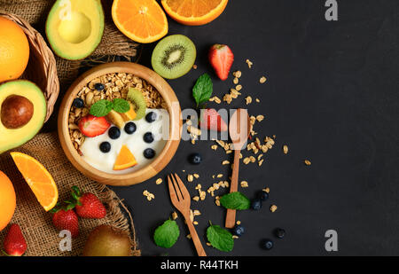 Müsli und Joghurt mit Früchten in Holz Schüssel und Früchte auf schwarzen Steintisch, gesunde Ernährung Konzept Stockfoto