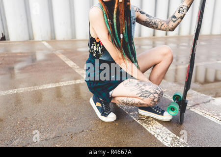 Junges Mädchen mit Tattoos und Dreadlocks sitzt mit einem Longboard auf dem Hintergrund der konkreten Struktur Stockfoto