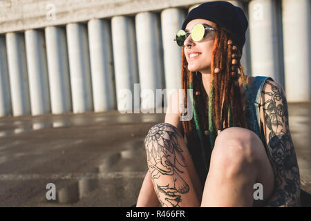 Junges Mädchen mit Tattoos und Dreadlocks in eine blaue Kappe sitzt auf einem longboard vor dem Hintergrund der konkreten Struktur Stockfoto