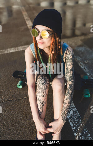 Junges Mädchen mit Tattoos und Dreadlocks in eine blaue Kappe sitzt auf einem longboard vor dem Hintergrund der konkreten Struktur Stockfoto