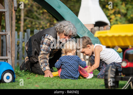 Großvater mit Enkel und Enkelin draußen spielen auf im Hinterhof sitzen auf Gras mit Spielzeug umgeben. Stockfoto