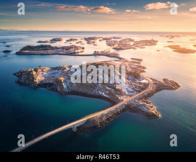 Luftaufnahme der Brücke über das Meer und die verschneiten Berge auf den Lofoten Inseln, Norwegen. Henningsvær bei Sonnenuntergang im Winter. Landschaft mit blauen Wasser, Himmel wi Stockfoto