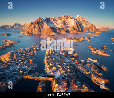 Luftaufnahme von henningsvær bei Sonnenuntergang auf den Lofoten Inseln, Norwegen. Winterlandschaft mit Gebäuden, schneebedeckten Bergen, blauen Meer. Blick von oben auf die Häuser, Boot Stockfoto