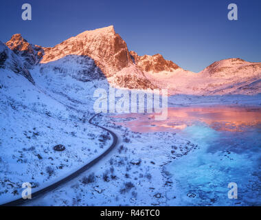 Luftaufnahme der kurvenreichen Straße, schneebedeckte Berge und blauer Himmel im Wasser bei Sonnenuntergang wider. Winterlandschaft mit Blick auf das Meer, die eisigen Küste, verschneite Felsen, r Stockfoto