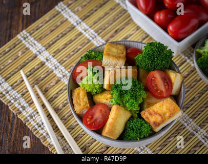 Gegrillter Tofu mit Brokkoli und Tomaten in Weiß Schüssel auf Bambus Pad mit Stäbchen auf der linken Seite. Stockfoto