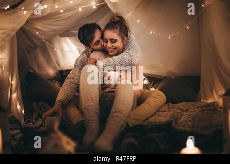Mann seine Freundin von hinten sitzen auf dem Bett in der Nacht und sie zu küssen. Ihre Frau sitzt auf dem Bett mit einer Tasse Kaffee mit ihrem Mann küssen Stockfoto