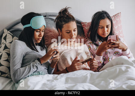 Drei Mädchen sitzt auf dem Bett und die Verwendung von Handys. Mädchen während einer Übernachtung auf dem Bett sitzen zusammen auf der Suche auf ihre Handys. Stockfoto